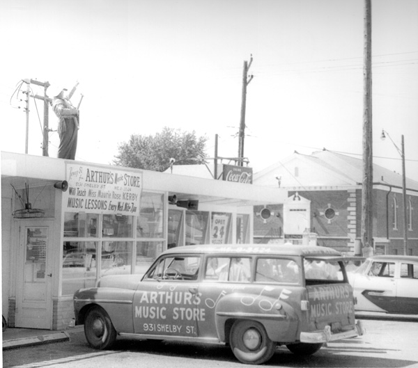 Arthur’s Music Store employee Kenny Si gives a rooftop guitar lesson to a pole-sitter using the 1958 Gibson Flying V later known as “Amos.” The store’s delivery vehicle is parked below.